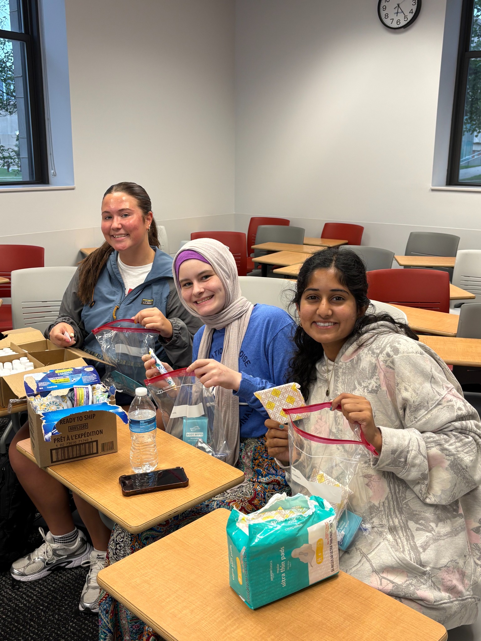 Three BAHT members holding hygiene bags while packing toiletries and menstrual products at a classroom table.
